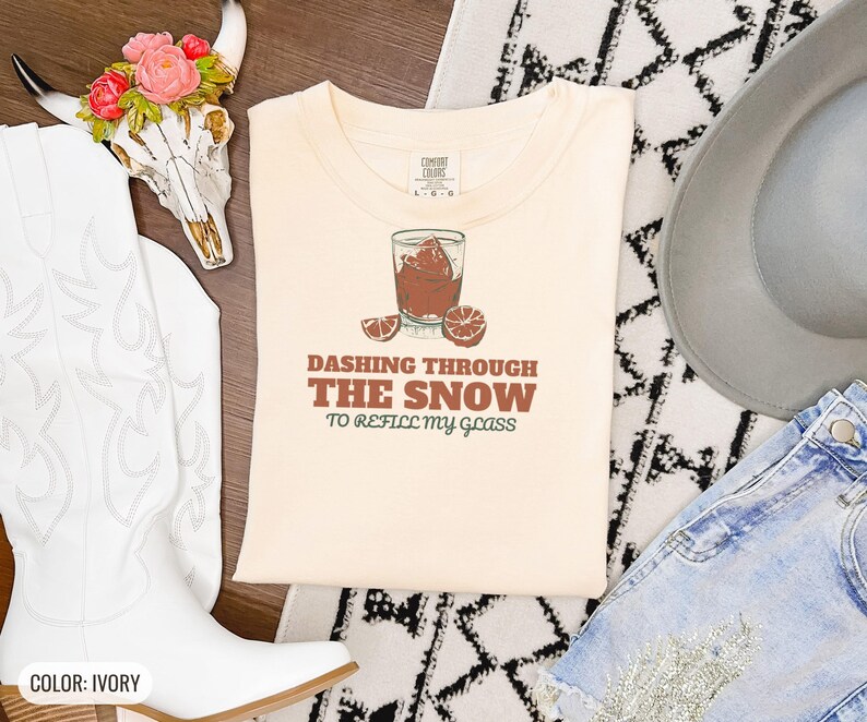 Ivory Comfort Colors t-shirt printed with the words Dashing Through The Snow To Refill My Glass and a drawing of a heavy crystal whiskey tumbler with two floating ice cubes beside a sprig of pine needles, folded flat lay