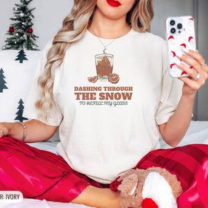 Ivory Comfort Colors t-shirt printed with the words Dashing Through The Snow To Refill My Glass and a drawing of a heavy crystal whiskey tumbler with two floating ice cubes beside a sprig of pine needles, worn by a woman
