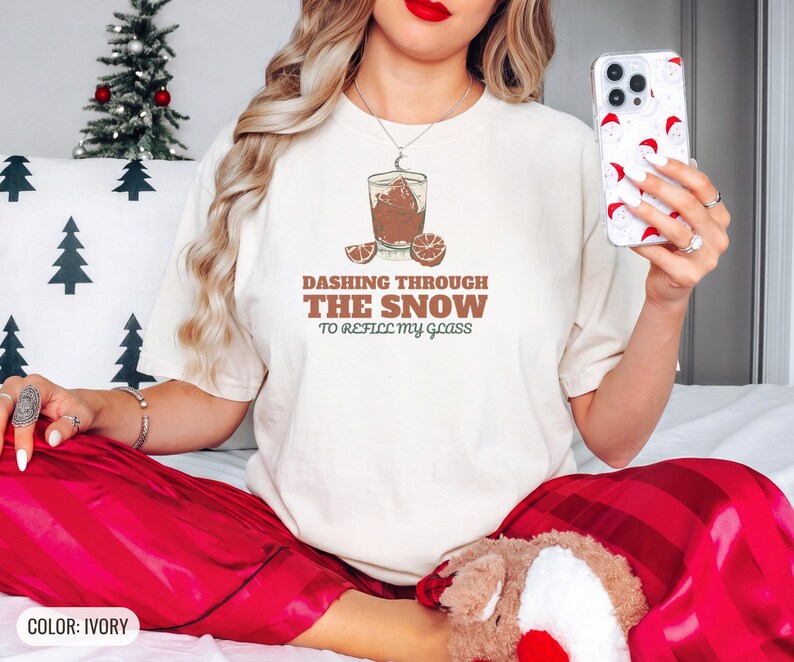 Ivory Comfort Colors t-shirt printed with the words Dashing Through The Snow To Refill My Glass and a drawing of a heavy crystal whiskey tumbler with two floating ice cubes beside a sprig of pine needles, worn by a woman