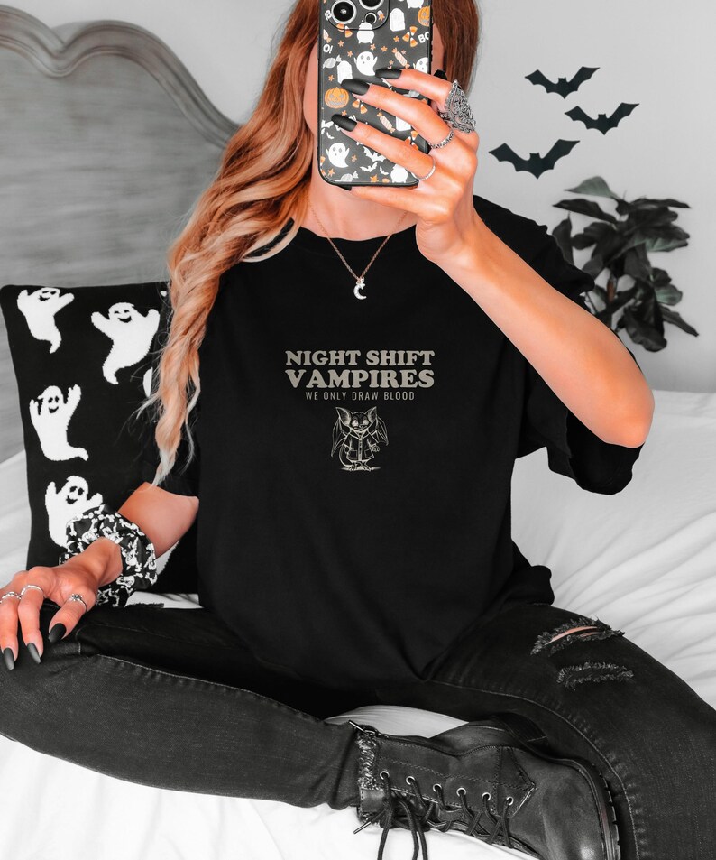 Black Comfort Colors t-shirt printed with the words Night Shift Vampires We Only Draw Blood and a drawing of a cute cartoon vampire bat wearing a tiny nurse scrub top, worn by a woman