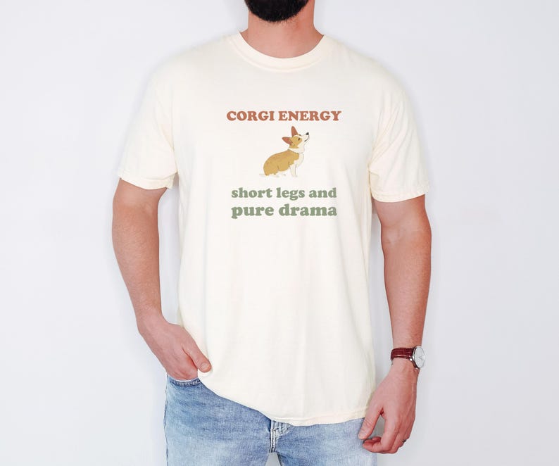 Ivory Comfort Colors t-shirt printed with the words Corgi Energy Short Legs And Pure Drama and a drawing of a corgi seen from behind with a fluffy heart-shaped butt, looking back over its shoulder, worn by a man
