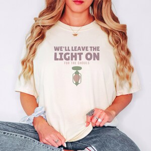 Ivory Comfort Colors t-shirt printed with the words We'Ll Leave The Light On For The Ghouls and a drawing of an antique black metal porch light fixture with a glowing yellow bulb, a tiny spiderweb in, worn by a woman