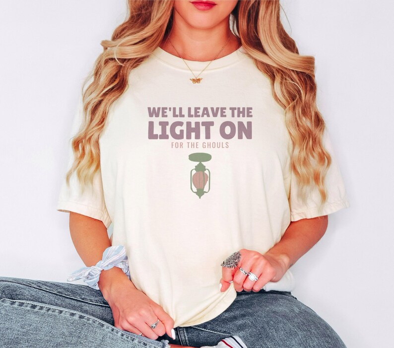 Ivory Comfort Colors t-shirt printed with the words We'Ll Leave The Light On For The Ghouls and a drawing of an antique black metal porch light fixture with a glowing yellow bulb, a tiny spiderweb in, worn by a woman