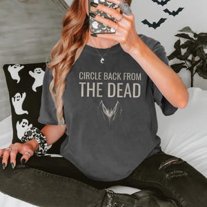 Pepper Comfort Colors t-shirt printed with the words Circle Back From The Dead and a drawing of a vintage silver paperclip bent into the shape of a bat, worn by a woman