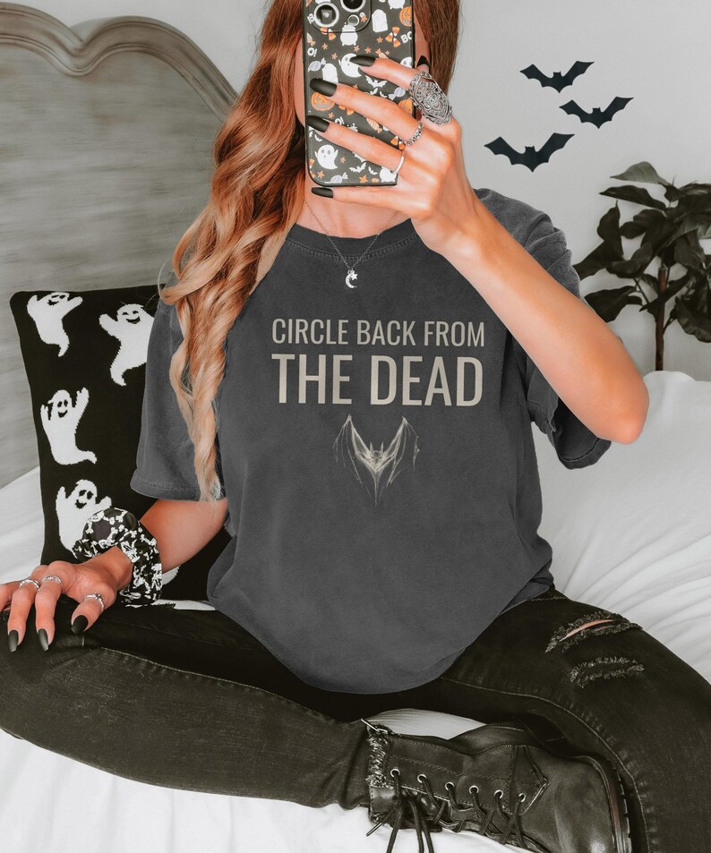 Pepper Comfort Colors t-shirt printed with the words Circle Back From The Dead and a drawing of a vintage silver paperclip bent into the shape of a bat, worn by a woman