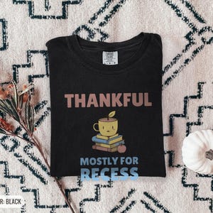 Black Comfort Colors t-shirt printed with the words Thankful Mostly For Recess and a drawing of a stack of retro school books topped with a glossy red apple and a steaming ceramic coffee, worn by a woman