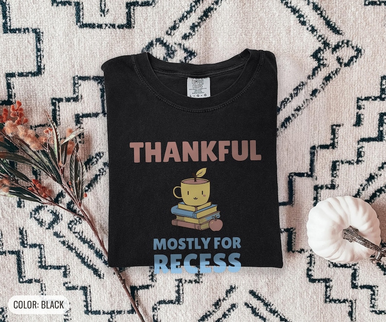 Black Comfort Colors t-shirt printed with the words Thankful Mostly For Recess and a drawing of a stack of retro school books topped with a glossy red apple and a steaming ceramic coffee, worn by a woman