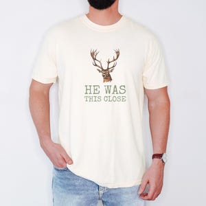 Ivory Comfort Colors t-shirt printed with the words He Was This Close and a drawing of a single large twelve-point deer antler shed resting on dry oak leaves, worn by a man