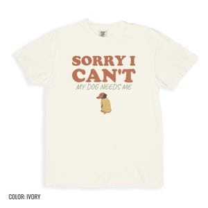 Ivory Comfort Colors t-shirt printed with the words Sorry I Can'T My Dog Needs Me and a drawing of a sleeping dachshund wearing a patterned knit sweater, folded flat lay