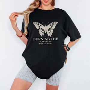 Black Comfort Colors t-shirt printed with the words Burning The Midnight Oil With The Moths and a drawing of a cecropia moth with feathery antennae, worn by a woman