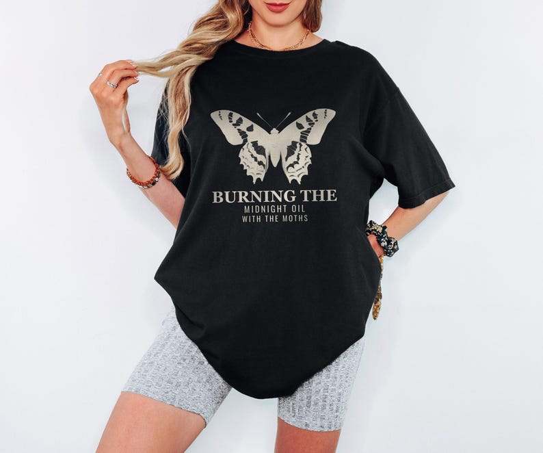 Black Comfort Colors t-shirt printed with the words Burning The Midnight Oil With The Moths and a drawing of a cecropia moth with feathery antennae, worn by a woman