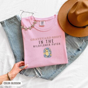 Blossom Comfort Colors t-shirt printed with the words Graveyard Shift In The Wildflower Patch and a drawing of a cute chubby ghost wearing a flower crown and holding a single violet, folded flat lay