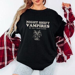Black Comfort Colors t-shirt printed with the words Night Shift Vampires We Only Draw Blood and a drawing of a cute cartoon vampire bat wearing a tiny nurse scrub top, worn by a woman