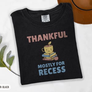 Black Comfort Colors t-shirt printed with the words Thankful Mostly For Recess and a drawing of a stack of retro school books topped with a glossy red apple and a steaming ceramic coffee, folded flat lay