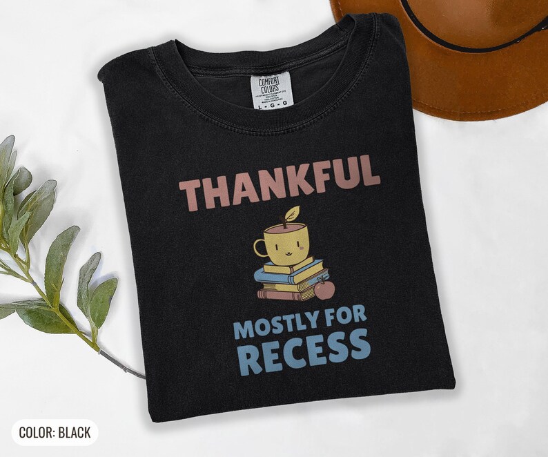Black Comfort Colors t-shirt printed with the words Thankful Mostly For Recess and a drawing of a stack of retro school books topped with a glossy red apple and a steaming ceramic coffee, folded flat lay