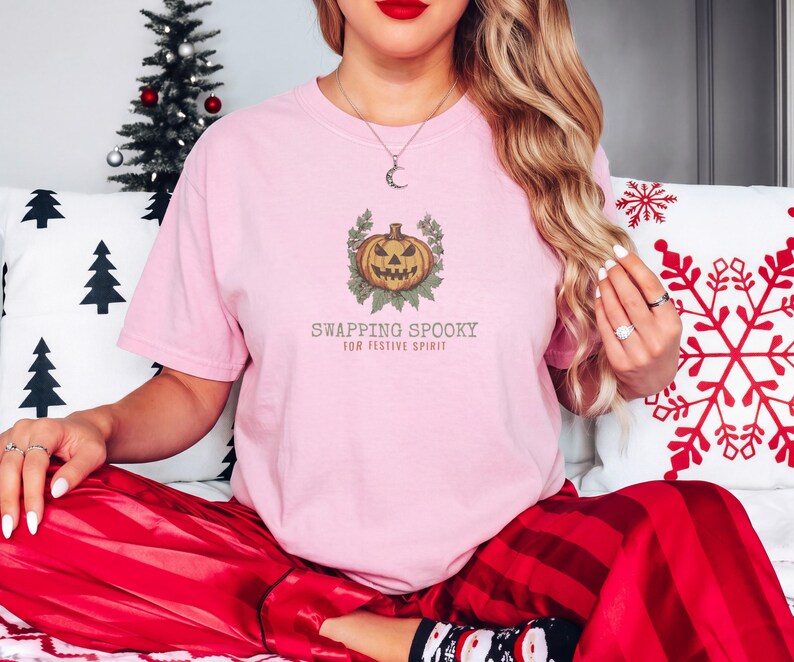 Blossom Comfort Colors t-shirt printed with the words Swapping Spooky For Festive Spirit and a drawing of a spooky jack-o-lantern pumpkin alongside a festive holiday wreath, worn by a woman