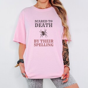 Blossom Comfort Colors t-shirt printed with the words Scared To Death By Their Spelling and a drawing of a highly detailed, crisp linocut spider sitting on top of a red-graded spelling test paper, worn by a woman