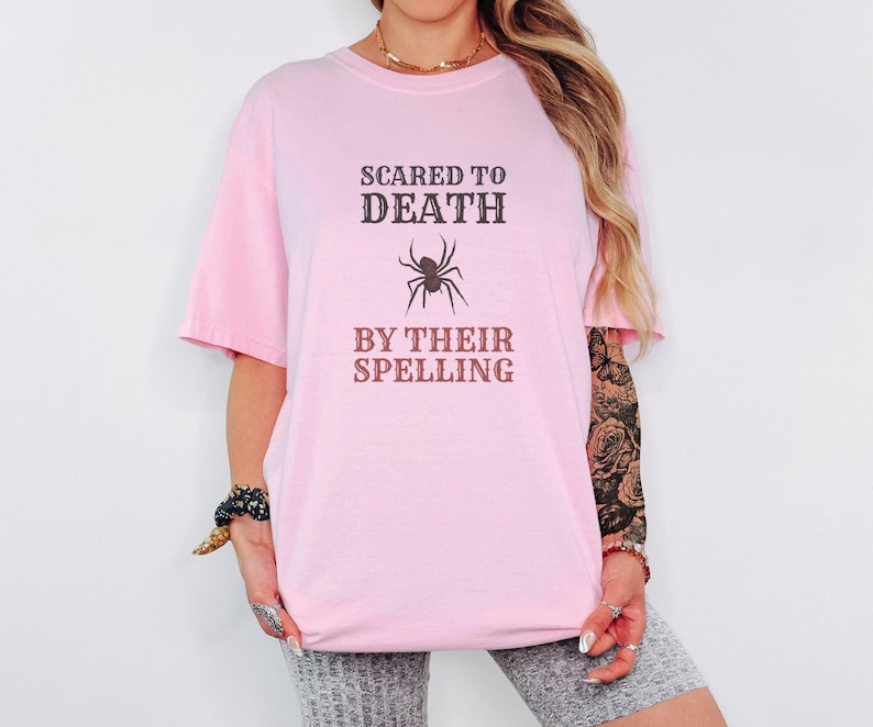 Blossom Comfort Colors t-shirt printed with the words Scared To Death By Their Spelling and a drawing of a highly detailed, crisp linocut spider sitting on top of a red-graded spelling test paper, worn by a woman