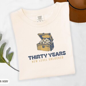 Ivory Comfort Colors t-shirt printed with the words Thirty Years New Level Unlocked and a drawing of a pixel-art treasure chest bursting open with light, folded flat lay