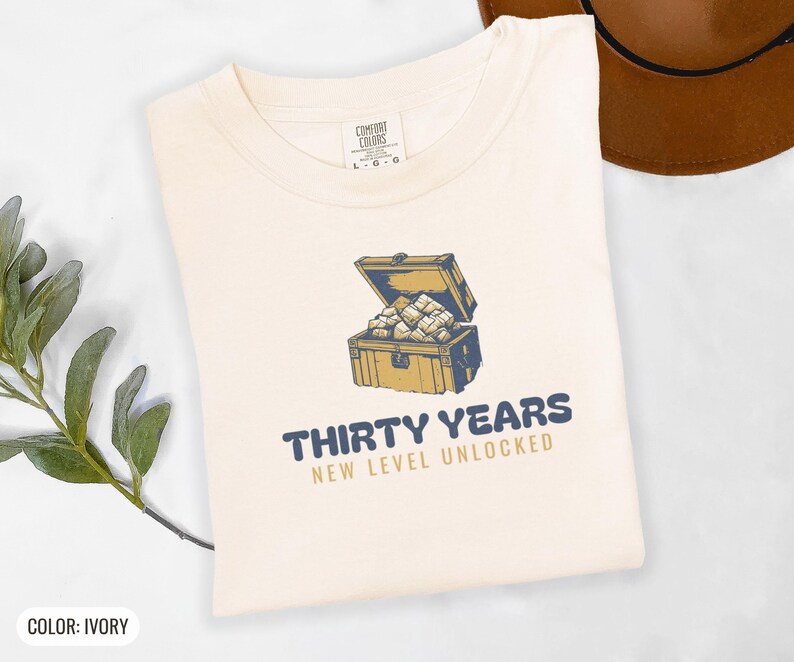 Ivory Comfort Colors t-shirt printed with the words Thirty Years New Level Unlocked and a drawing of a pixel-art treasure chest bursting open with light, folded flat lay