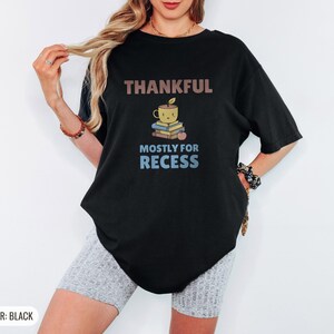Black Comfort Colors t-shirt printed with the words Thankful Mostly For Recess and a drawing of a stack of retro school books topped with a glossy red apple and a steaming ceramic coffee, worn by a woman