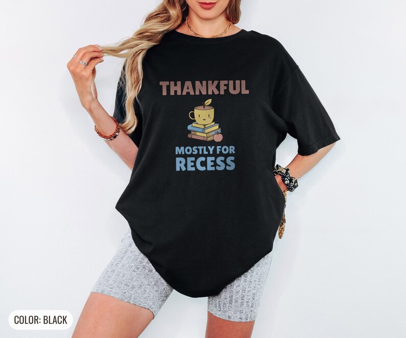 Black Comfort Colors t-shirt printed with the words Thankful Mostly For Recess and a drawing of a stack of retro school books topped with a glossy red apple and a steaming ceramic coffee, worn by a woman