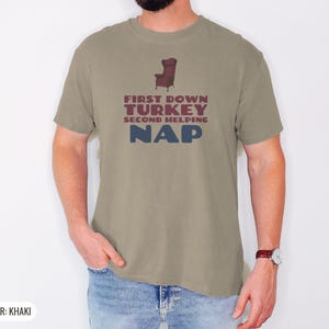 Khaki Comfort Colors t-shirt printed with the words First Down Turkey Second Helping Nap and a drawing of a vintage brown recliner with a footrest extended outward, worn by a man