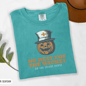 Seafoam Comfort Colors t-shirt printed with the words No Rest For The Wicked Or The Triage Nurse and a drawing of a grinning jack-o'-lantern wearing a classic nurse's cap with a red cross, folded flat lay
