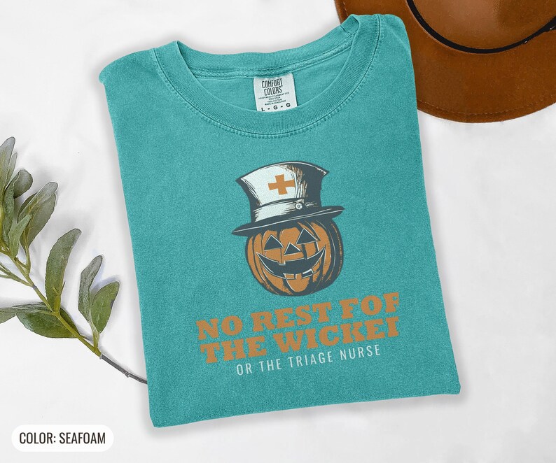 Seafoam Comfort Colors t-shirt printed with the words No Rest For The Wicked Or The Triage Nurse and a drawing of a grinning jack-o'-lantern wearing a classic nurse's cap with a red cross, folded flat lay