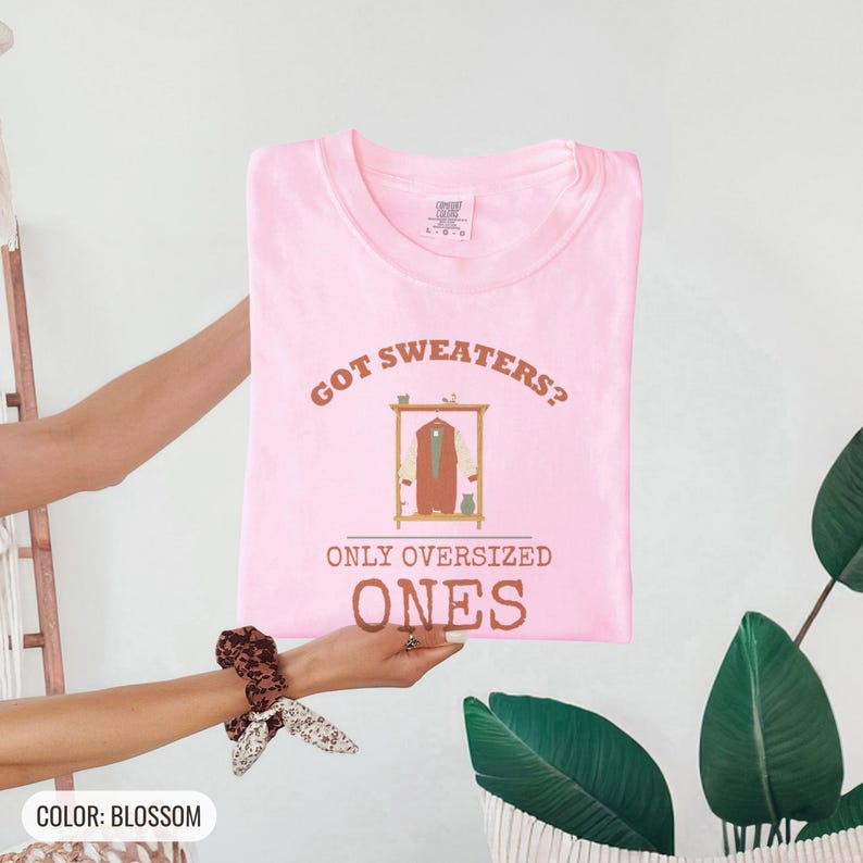 Blossom Comfort Colors t-shirt printed with the words Got Sweaters? Only Oversized Ones and a drawing of a knitted wool cardigan hanging from a simple wooden clothes hanger, folded flat lay