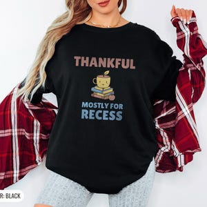 Black Comfort Colors t-shirt printed with the words Thankful Mostly For Recess and a drawing of a stack of retro school books topped with a glossy red apple and a steaming ceramic coffee, worn by a woman