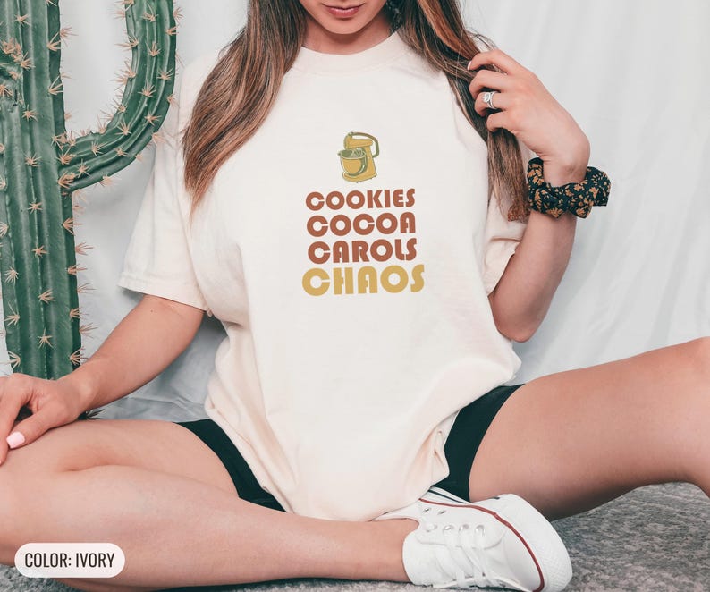Ivory Comfort Colors t-shirt printed with the words Cookies Cocoa Carols Chaos and a drawing of a retro metal hand mixer with steel beaters, worn by a woman