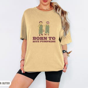 Butter Comfort Colors t-shirt printed with the words Born To Bite Pumpkins and a drawing of pair of corduroy toddler overalls splattered with pumpkin pulp, worn by a woman