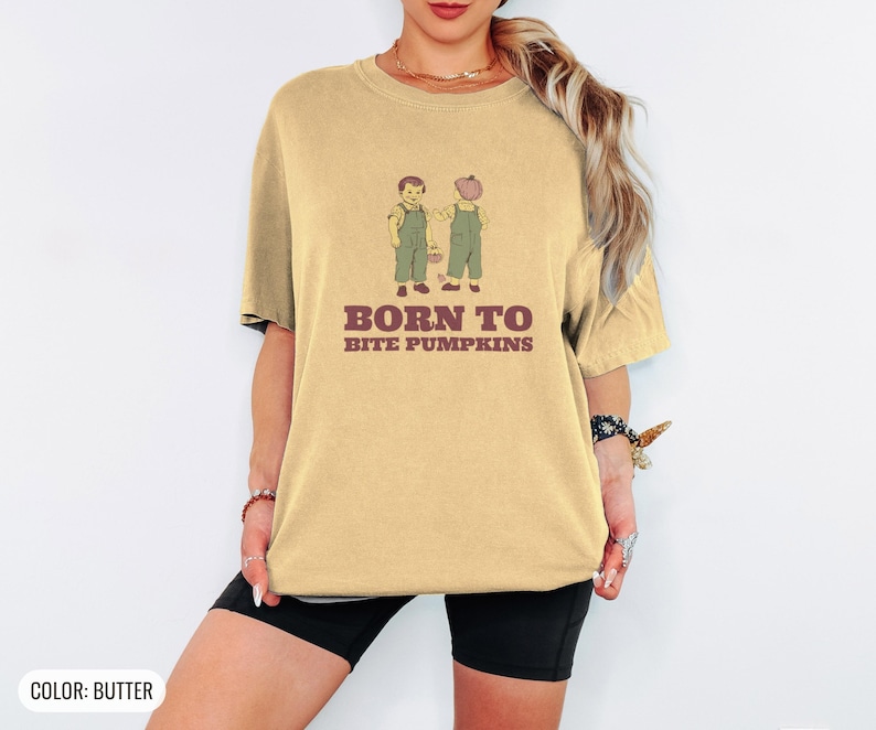 Butter Comfort Colors t-shirt printed with the words Born To Bite Pumpkins and a drawing of pair of corduroy toddler overalls splattered with pumpkin pulp, worn by a woman