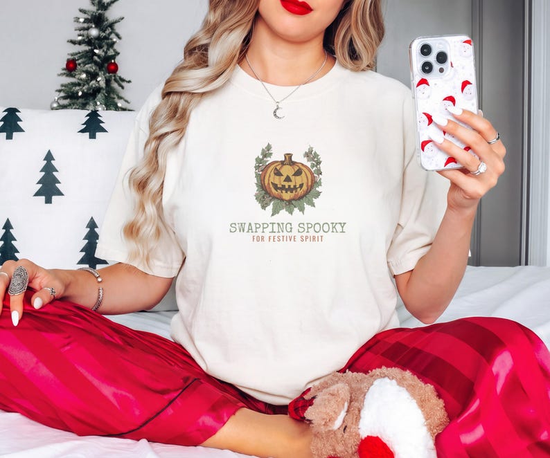 Ivory Comfort Colors t-shirt printed with the words Swapping Spooky For Festive Spirit and a drawing of a spooky jack-o-lantern pumpkin alongside a festive holiday wreath, worn by a woman