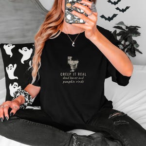 Black Comfort Colors t-shirt printed with the words Creep It Real Dead Leaves And Pumpkin Rinds and a drawing of a grinning skull with black sunglasses, worn by a woman
