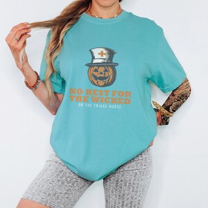 Seafoam Comfort Colors t-shirt printed with the words No Rest For The Wicked Or The Triage Nurse and a drawing of a grinning jack-o'-lantern wearing a classic nurse's cap with a red cross, worn by a woman