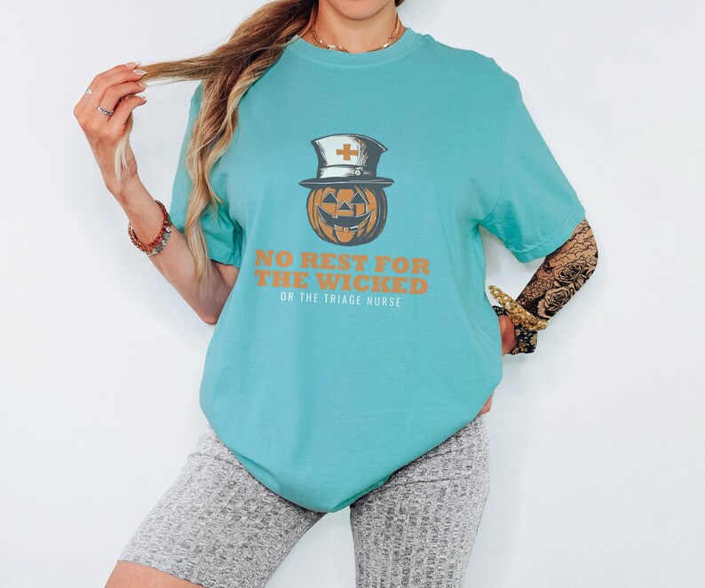 Seafoam Comfort Colors t-shirt printed with the words No Rest For The Wicked Or The Triage Nurse and a drawing of a grinning jack-o'-lantern wearing a classic nurse's cap with a red cross, worn by a woman