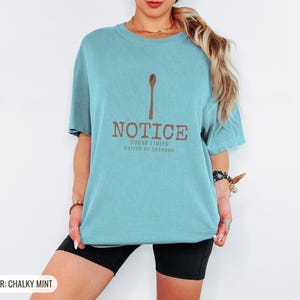 Chalky Mint Comfort Colors t-shirt printed with the words Notice Sugar Limits Waived By Grandma and a drawing of a painted wooden spoon crossed over a cinnamon stick bundle, worn by a woman