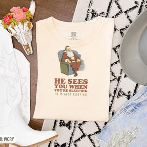 Ivory Comfort Colors t-shirt printed with the words He Sees You When You'Re Sleeping He Is Also Sleeping and a drawing of santa Claus in unbuttoned flannel asleep on a patterned sofa beside a drowsy elf wearing a, folded flat lay