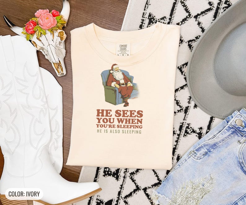Ivory Comfort Colors t-shirt printed with the words He Sees You When You'Re Sleeping He Is Also Sleeping and a drawing of santa Claus in unbuttoned flannel asleep on a patterned sofa beside a drowsy elf wearing a, folded flat lay