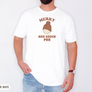 White Comfort Colors t-shirt printed with the words Merry And Under Par and a drawing of a dimpled golf ball wearing a tiny red and white striped knit winter beanie with a green p, worn by a man