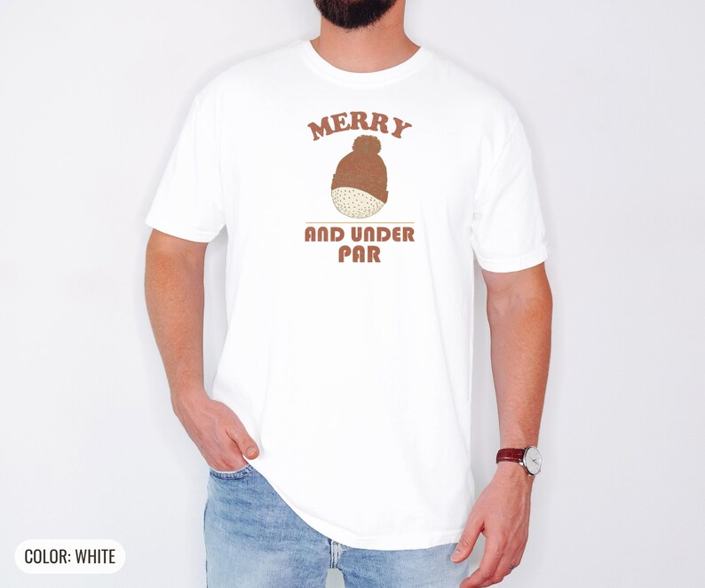 White Comfort Colors t-shirt printed with the words Merry And Under Par and a drawing of a dimpled golf ball wearing a tiny red and white striped knit winter beanie with a green p, worn by a man