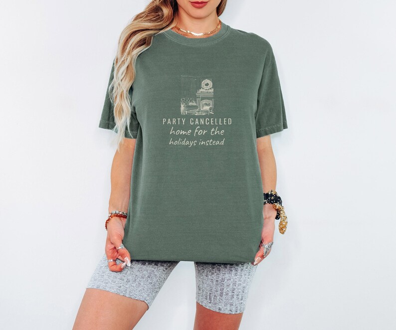 Moss Comfort Colors t-shirt printed with the words Party Cancelled Home For The Holidays Instead and a drawing of a cozy living room with a fireplace and Christmas decorations, a warm glow fills the space, worn by a woman