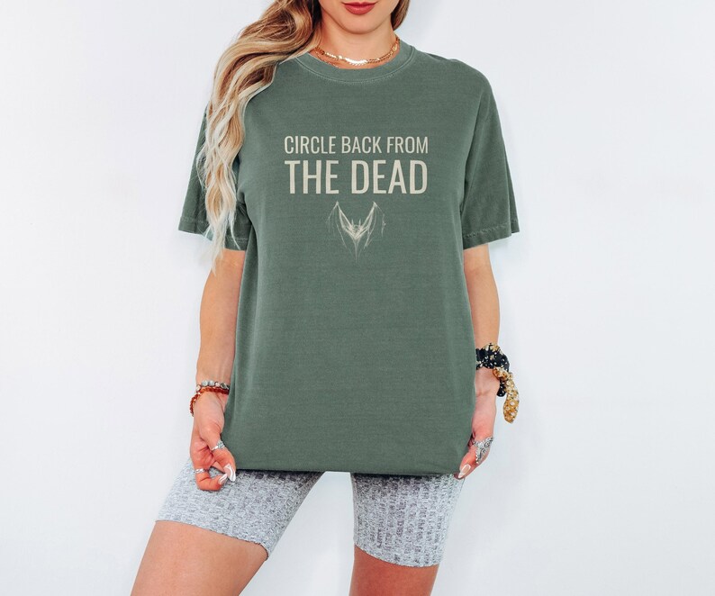 Moss Comfort Colors t-shirt printed with the words Circle Back From The Dead and a drawing of a vintage silver paperclip bent into the shape of a bat, worn by a woman