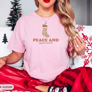 Blossom Comfort Colors t-shirt printed with the words Peace And Grandkids and a drawing of a classic knitted wool christmas stocking stuffed with small toys, worn by a woman