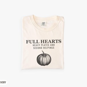 Ivory Comfort Colors t-shirt printed with the words Full Hearts Heavy Plates And Second Helpings and a drawing of a pumpkin surrounded by fall leaves and small acorns, folded flat lay
