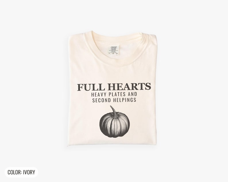 Ivory Comfort Colors t-shirt printed with the words Full Hearts Heavy Plates And Second Helpings and a drawing of a pumpkin surrounded by fall leaves and small acorns, folded flat lay