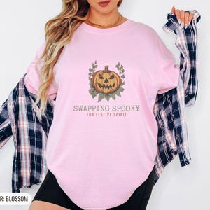 Blossom Comfort Colors t-shirt printed with the words Swapping Spooky For Festive Spirit and a drawing of a spooky jack-o-lantern pumpkin alongside a festive holiday wreath, worn by a woman