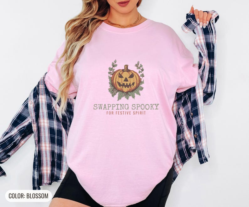 Blossom Comfort Colors t-shirt printed with the words Swapping Spooky For Festive Spirit and a drawing of a spooky jack-o-lantern pumpkin alongside a festive holiday wreath, worn by a woman